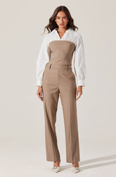 A woman is wearing a white, long-sleeved, collared shirt with vertical stripes. Over the shirt, she has on a strapless, beige tailored jumpsuit that fits snugly at the top and flows into wide-leg trousers. She is also wearing pointed white heels.